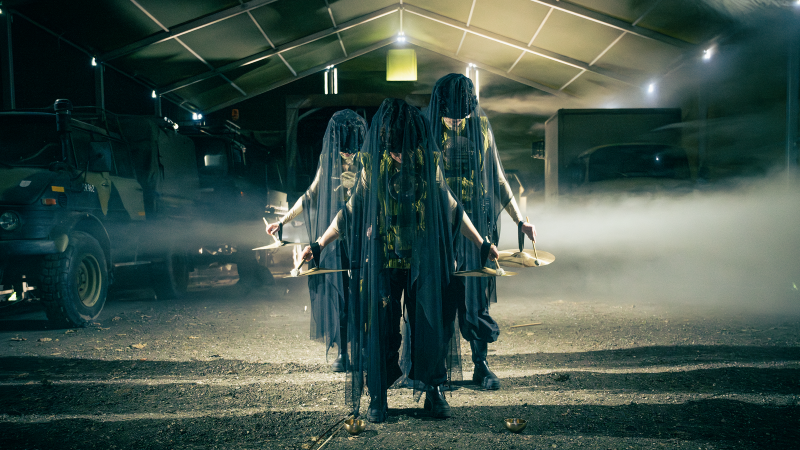 Tre personer i mørke kostumer, der dækker ansigterne, på scenen i en teaterforestilling