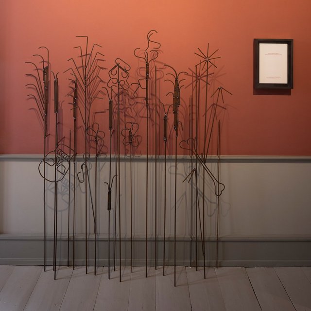 Et kunstværk, der ligner planter, foran en rød væg. 