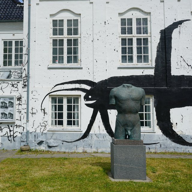 En statue foran en hvid bygning, hvorpå en stor sort figur er malet. 